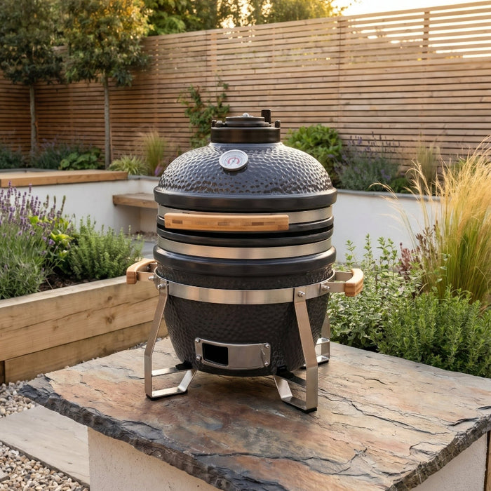 Outdoor setting with a large barbecue grill on a stone surface, surrounded by plants and wooden structures.